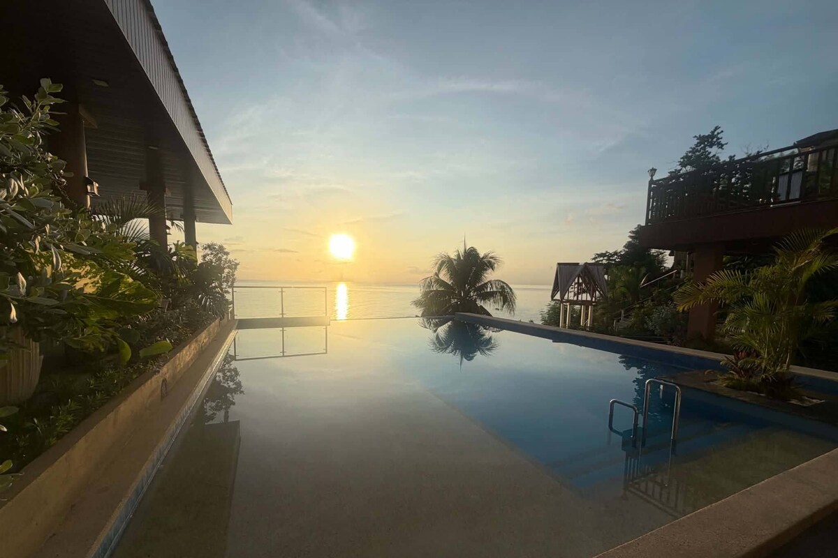 Visão nítida de uma piscina com o pôr do sol ao fundo, cercada por vegetação e edifícios.