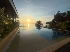 Visão nítida de uma piscina com o pôr do sol ao fundo, cercada por vegetação e edifícios.