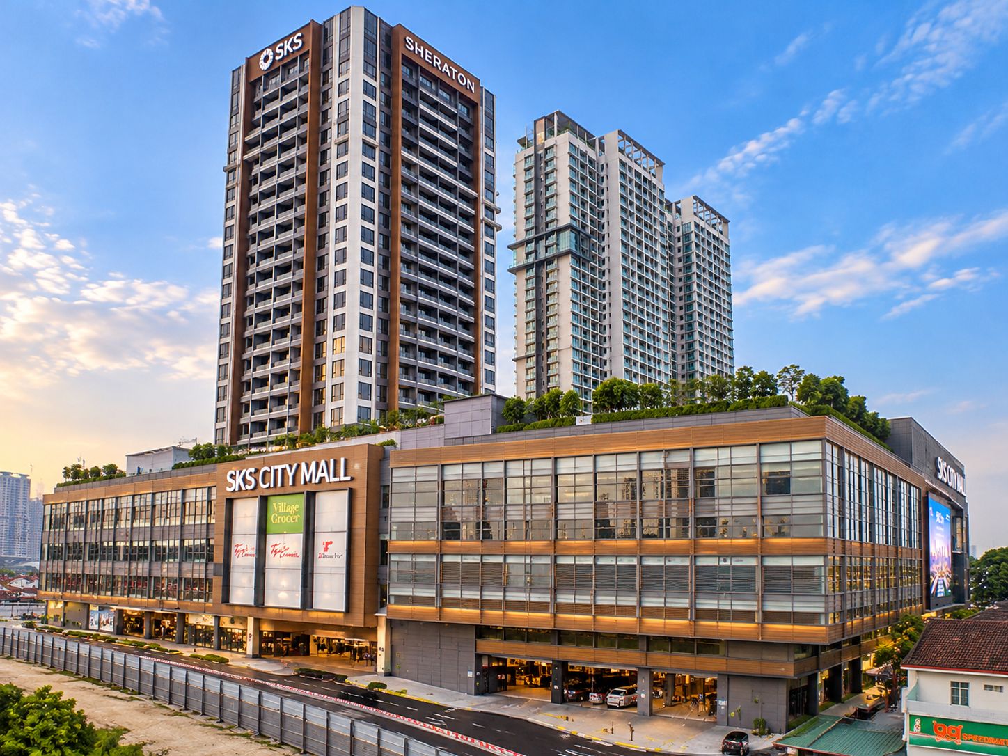 Un immeuble commercial moderne à plusieurs étages, doté de grandes fenêtres en verre et de panneaux indiquant « SKS CITY MALL » et « Village Grocer ».