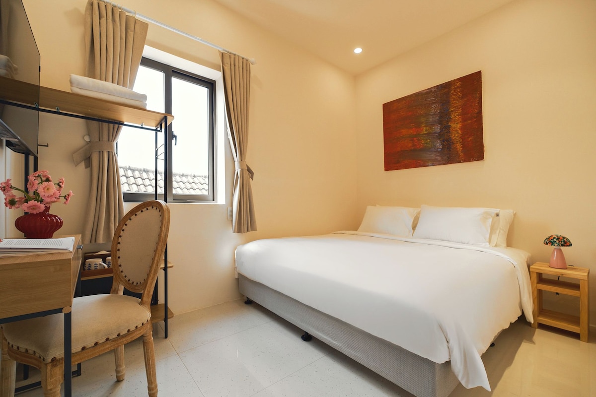 A bedroom with a bed, a desk, a chair, a window with curtains, a painting on the wall, and a nightstand with a lamp.