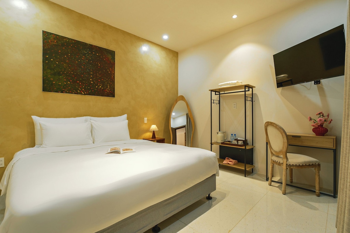 A hotel room with a large bed, a desk with a chair, a TV, a painting on the wall, and a mirror.