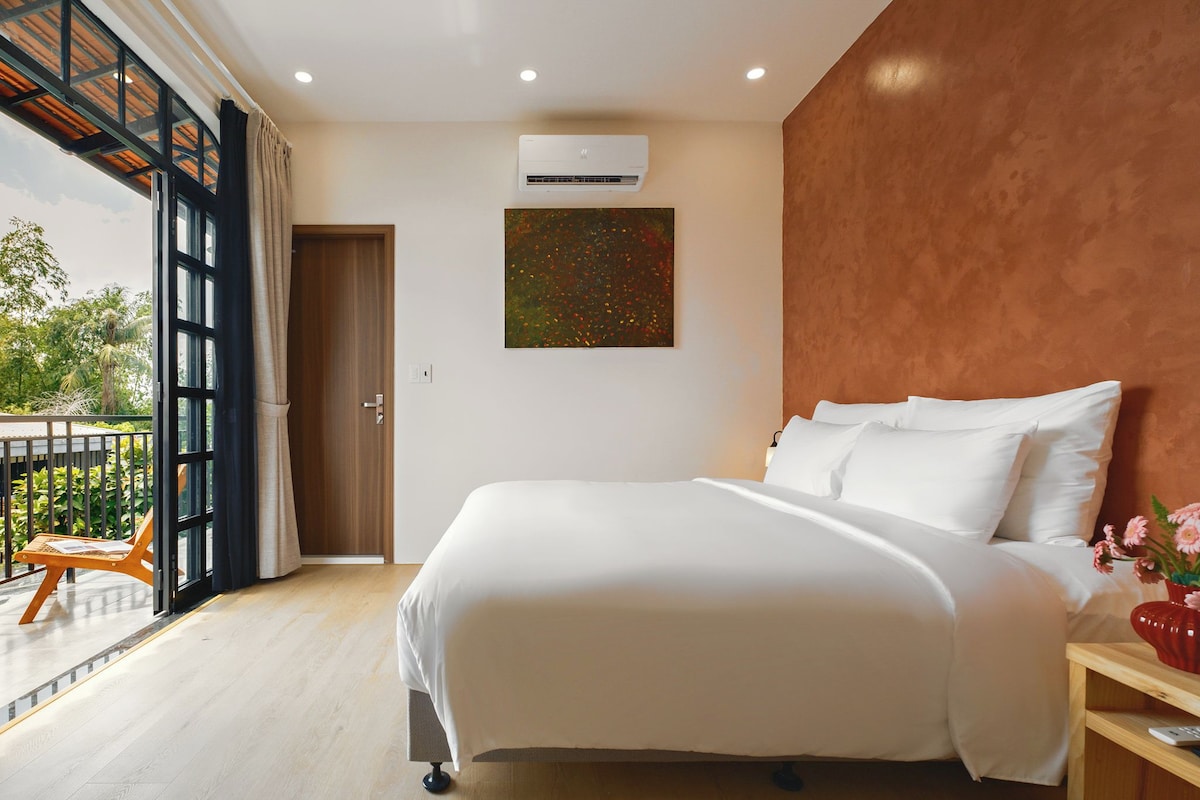 A bedroom with a large bed, an air conditioning unit, a painting, and a view of the outdoors.