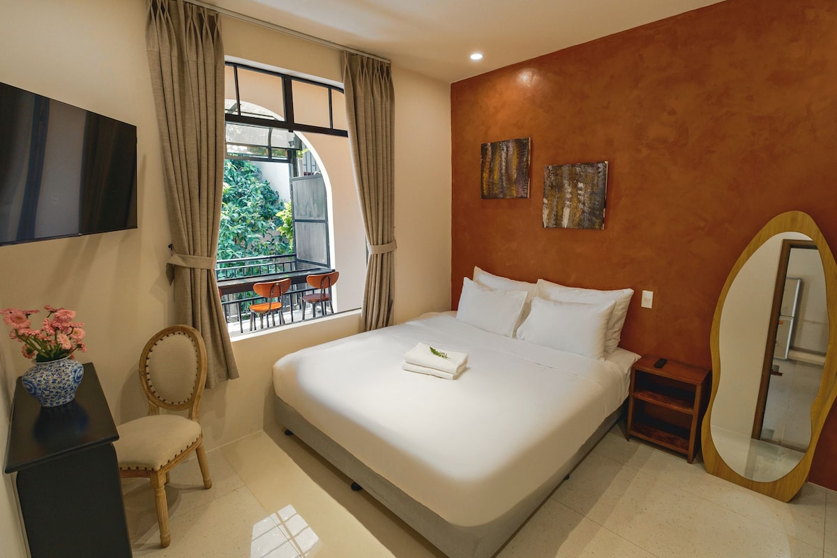A hotel room with a bed, a TV, a desk with a vase, a chair, a window with curtains, and a balcony.