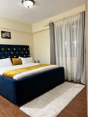 Ein Schlafzimmer mit einem Bett, einem Kopfteil, einem weißen Teppich und einem Fenster mit Vorhängen.