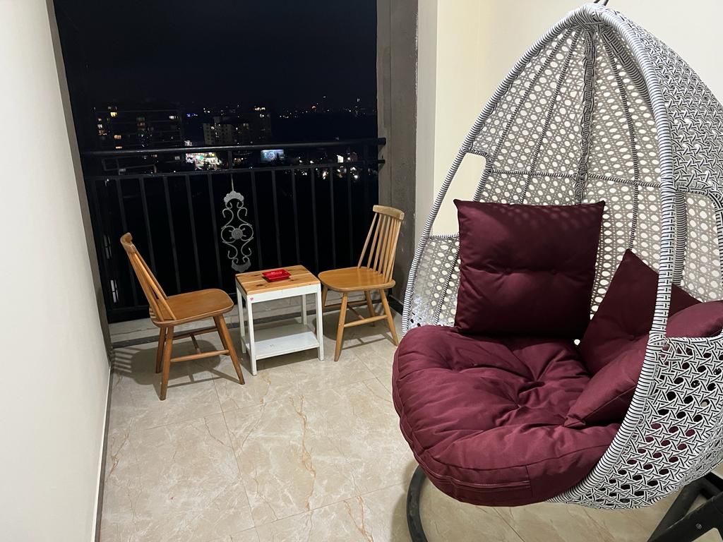 Ein Balkon mit einer Hängematte, zwei Holzstühlen, einem kleinen Tisch und einem roten Gegenstand, sowie ein Blick auf die Stadt bei Nacht.