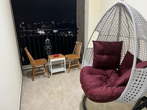 Ein Balkon mit einer Hängematte, zwei Holzstühlen, einem kleinen Tisch und einem roten Gegenstand, sowie ein Blick auf die Stadt bei Nacht.