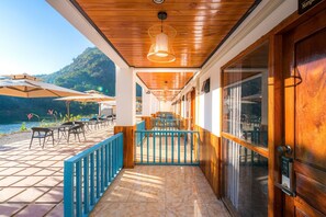 Een overdekt terras met tafels en stoelen, een houten plafond en uitzicht op een meer en bergen.