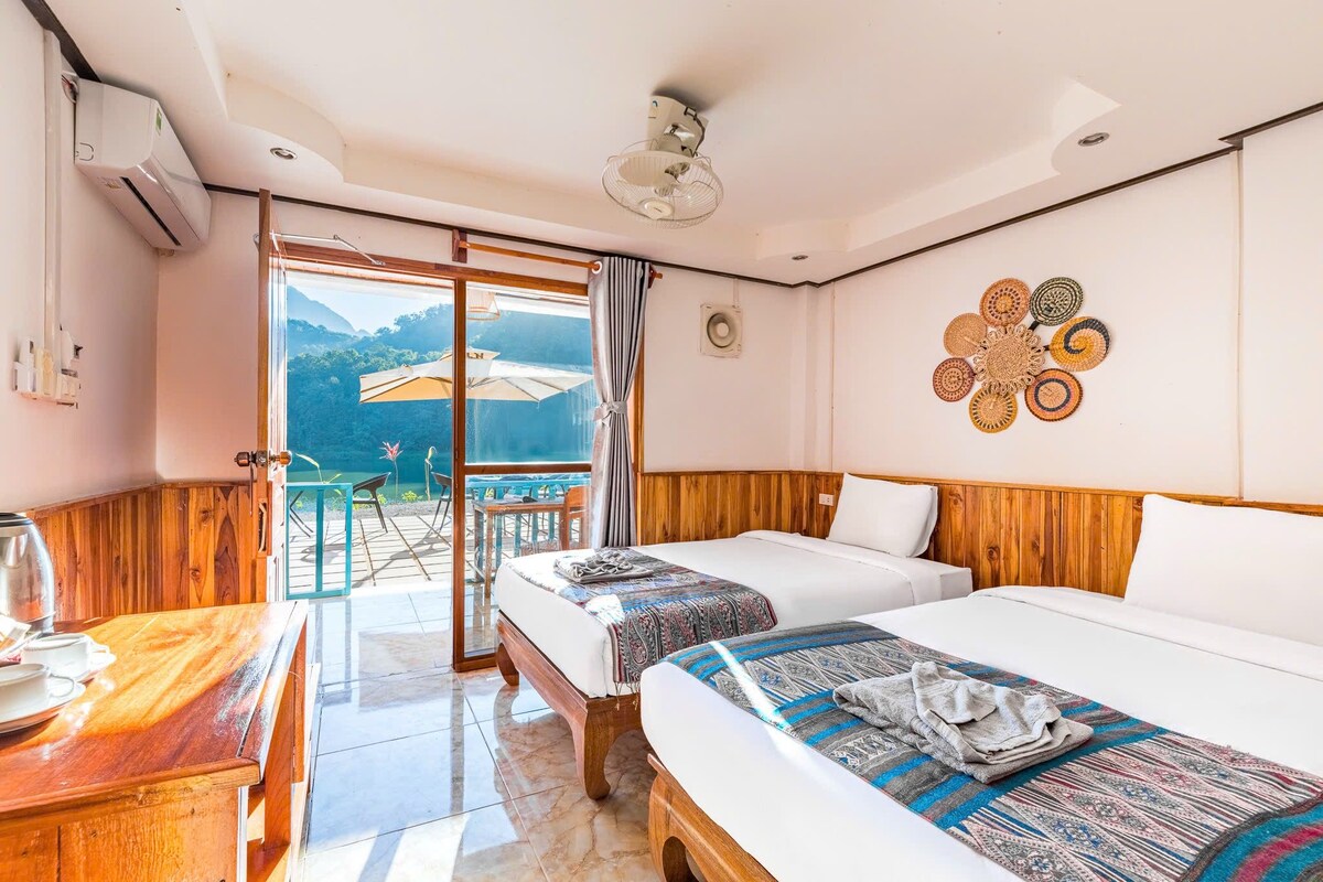 Een hotelkamer met twee bedden, een houten hoofdbord, een balkon met uitzicht en een tafel met waterkoker.