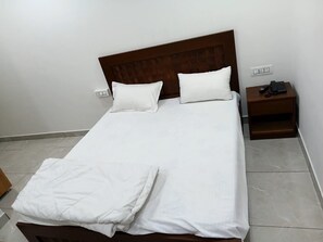 Ein ordentlich bezogenes Bett mit weißen Leinen und Kissen, ein hölzernes Kopfteil, ein Nachttisch mit Telefon und Fernbedienung sowie eine Steckdose an der Wand.