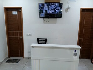 Ein Empfangsbereich mit einem weißen Schreibtisch, einem Stuhl und einem an der Wand befestigten Fernseher, der Überwachungsaufnahmen zeigt. Es befinden sich zwei Holztüren dort, eine mit der Aufschrift 'WASHROOM MALE FEMALE'.