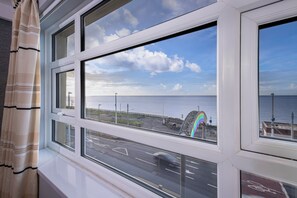 Une vue depuis une fenêtre donnant sur une route côtière, avec une structure aux couleurs de l’arc-en-ciel, un ciel dégagé et la mer à l’horizon.