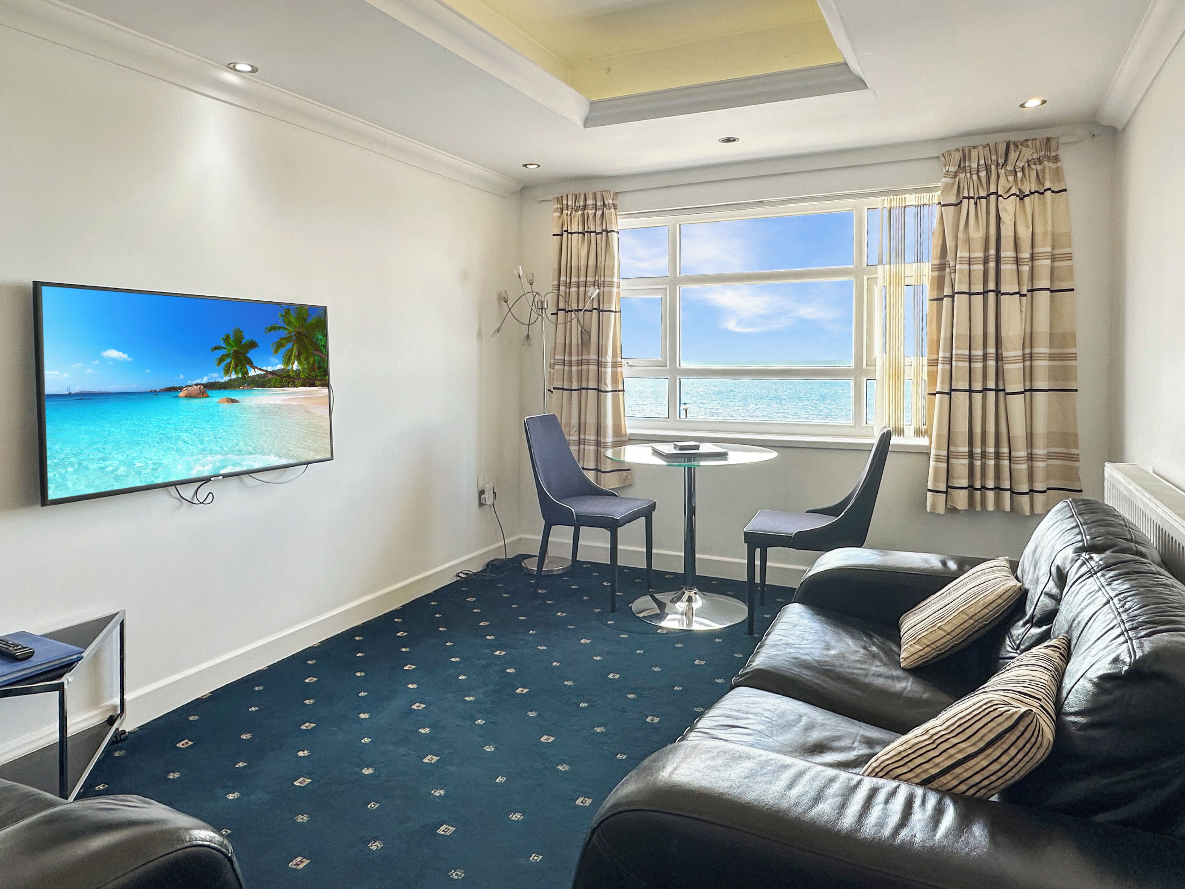 Quarto de hotel com uma televisão grande na parede, um sofá, uma mesinha e vista para o mar pela janela.
