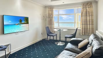 Quarto de hotel com uma televisão grande na parede, um sofá, uma mesinha e vista para o mar pela janela.