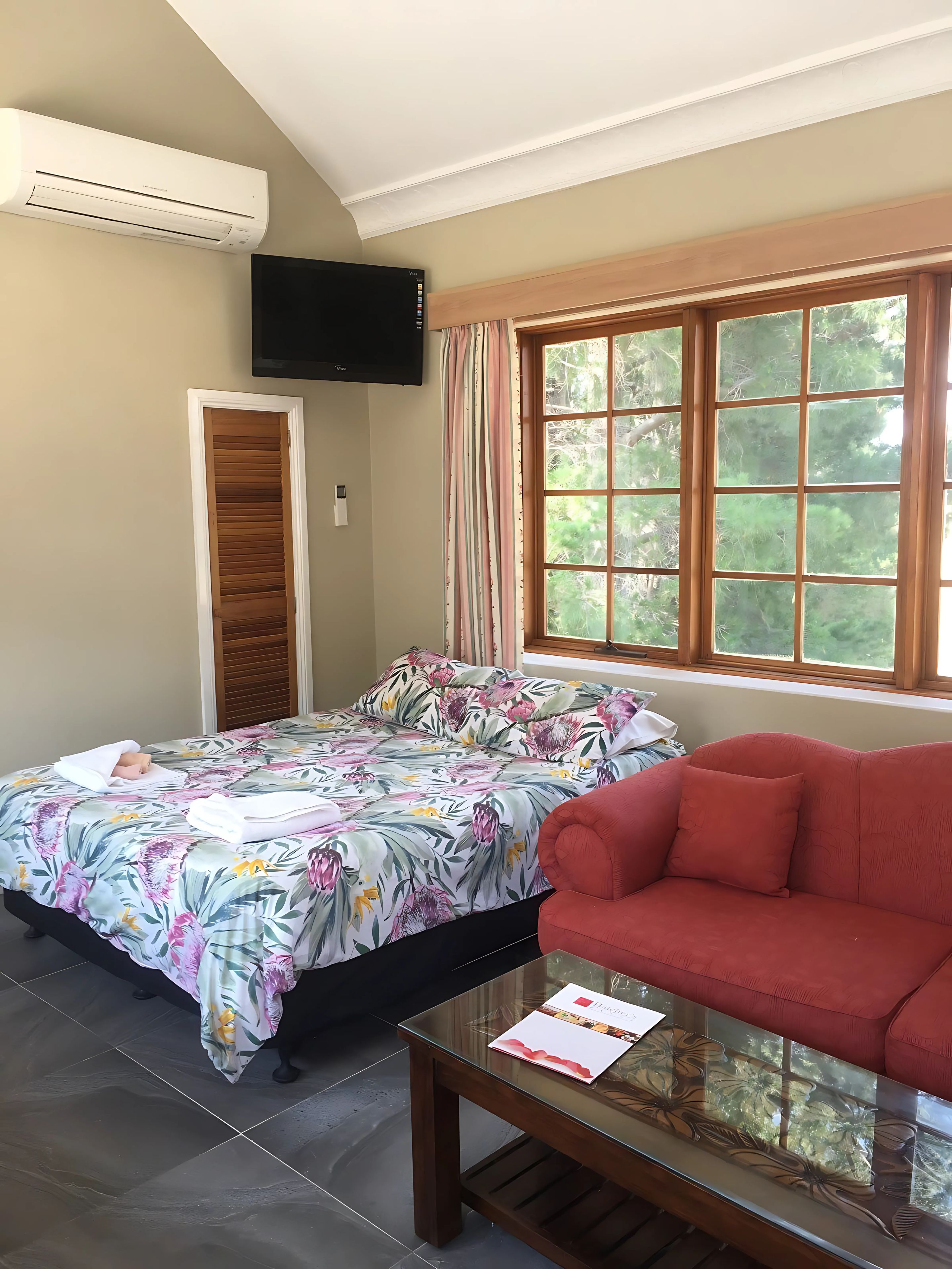 Ein Zimmer mit einem Bett, einem roten Sofa, einem Glastisch und einem Fenster mit Blick auf Bäume sowie einer Klimaanlage.