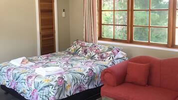 Ein Zimmer mit einem Bett, einem roten Sofa, einem Glastisch und einem Fenster mit Blick auf Bäume sowie einer Klimaanlage.