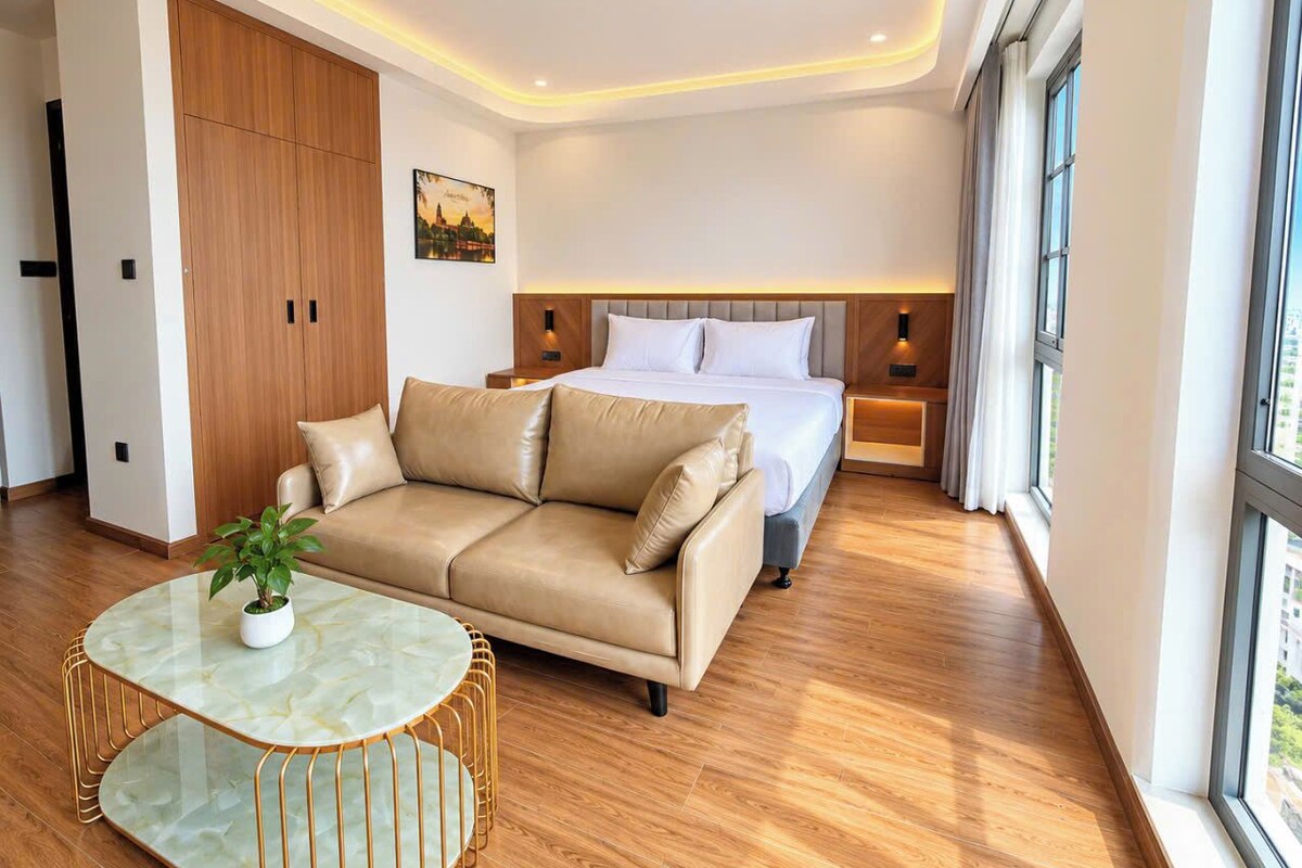 A modern hotel room with a wooden floor, a beige sofa, a round glass table with a plant, a bed with white linens, and a cityscape painting on the wall.