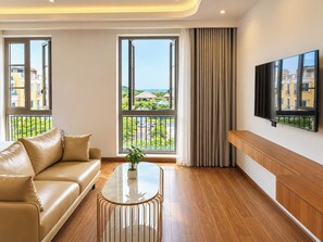 A modern living room with a sofa, a coffee table, a TV mounted on the wall, and large windows with a view of greenery and buildings. - JOYA HOTEL PHU QUOC (Phu Quoc)