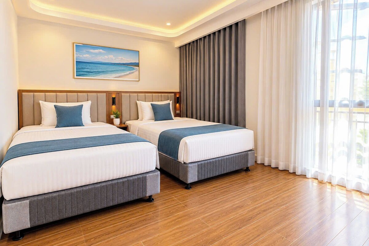 A hotel room with two beds, a large window with curtains, a painting on the wall, and a wooden floor.