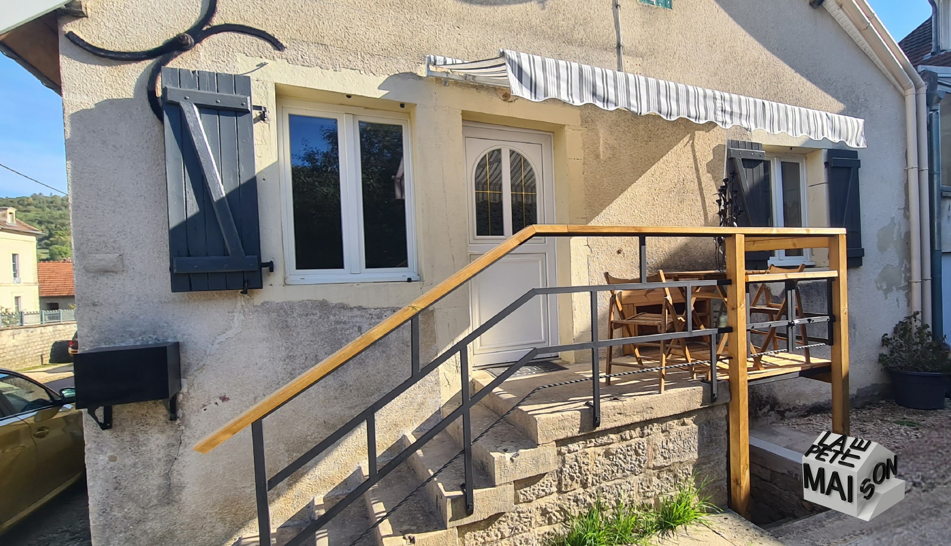 A building with a staircase leading to a wooden deck area, a sign reading 'LAW PETE MAISON,' and a window with a shutter.