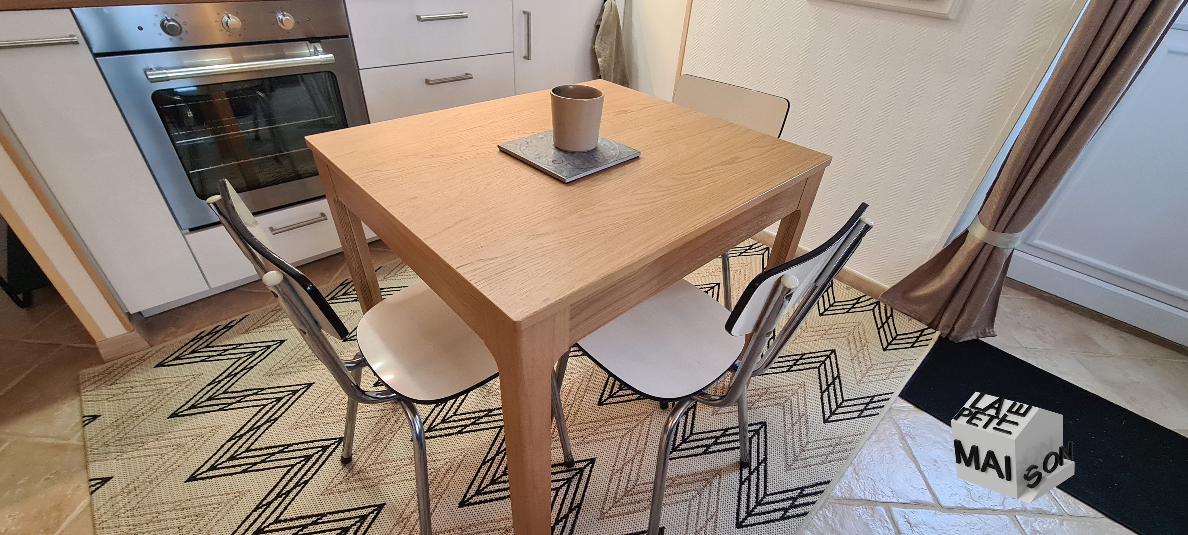 A small dining area with a wooden table, two chairs, a cup, and a doormat with 'LAW PETE MAISON' text.