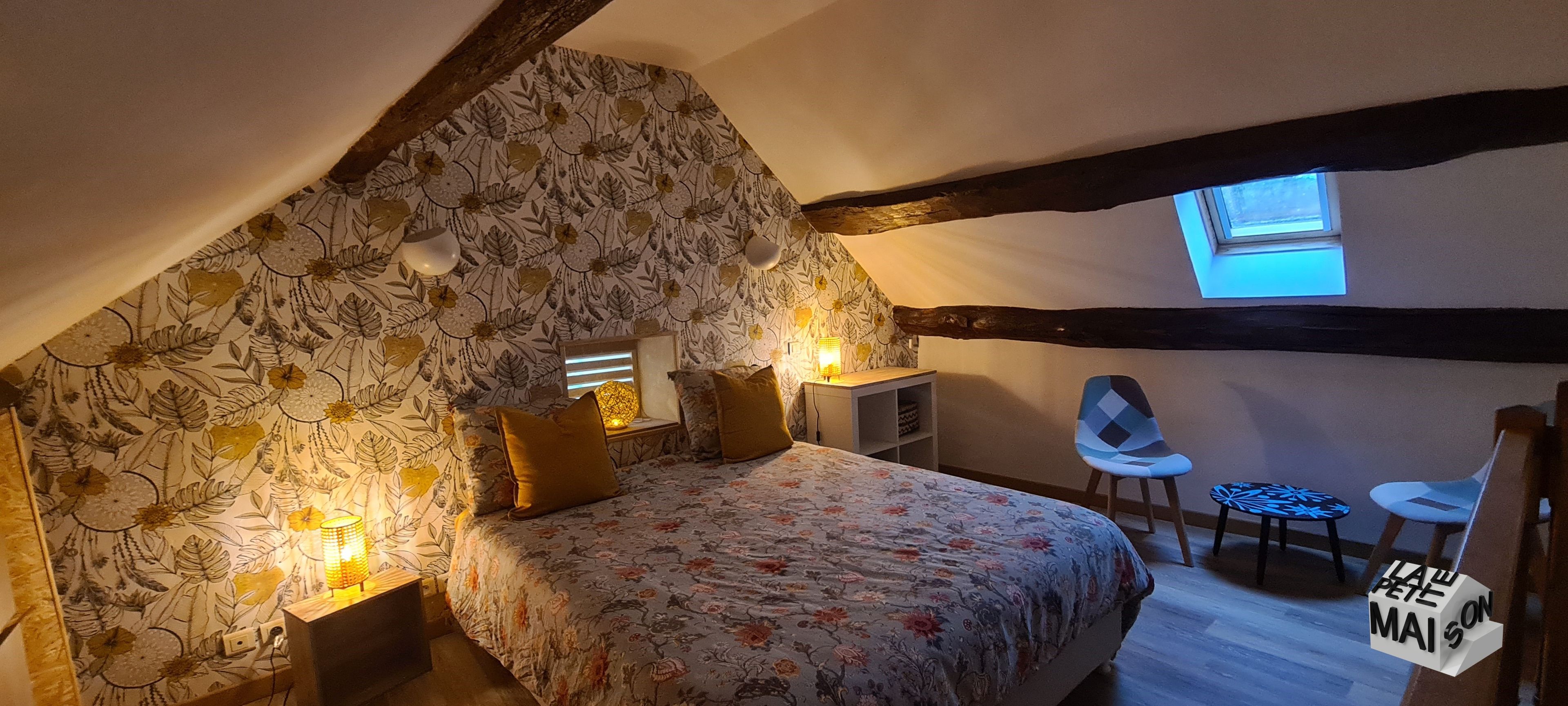 A bedroom with a sloped ceiling, a bed with floral bedding, two bedside lamps, a small table, a chair, and a window.