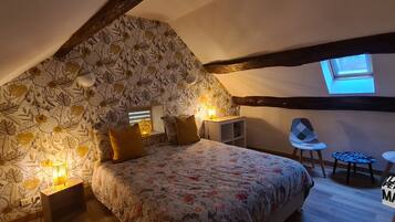 A bedroom with a sloped ceiling, a bed with floral bedding, two bedside lamps, a small table, a chair, and a window.