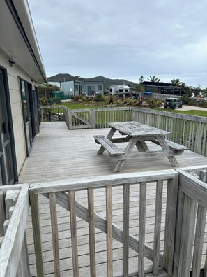 Eine Holzterrasse mit Picknicktisch, umgeben von einem Holzzaun, die zu einem Rasenbereich mit einem Haus und einem Wohnwagen im Hintergrund führt.