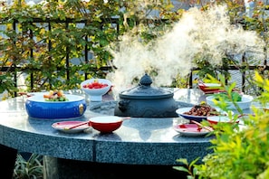 Une table ronde, sur laquelle sont disposés divers plats et un récipient d’où s’échappe de la vapeur, le tout entouré de verdure.