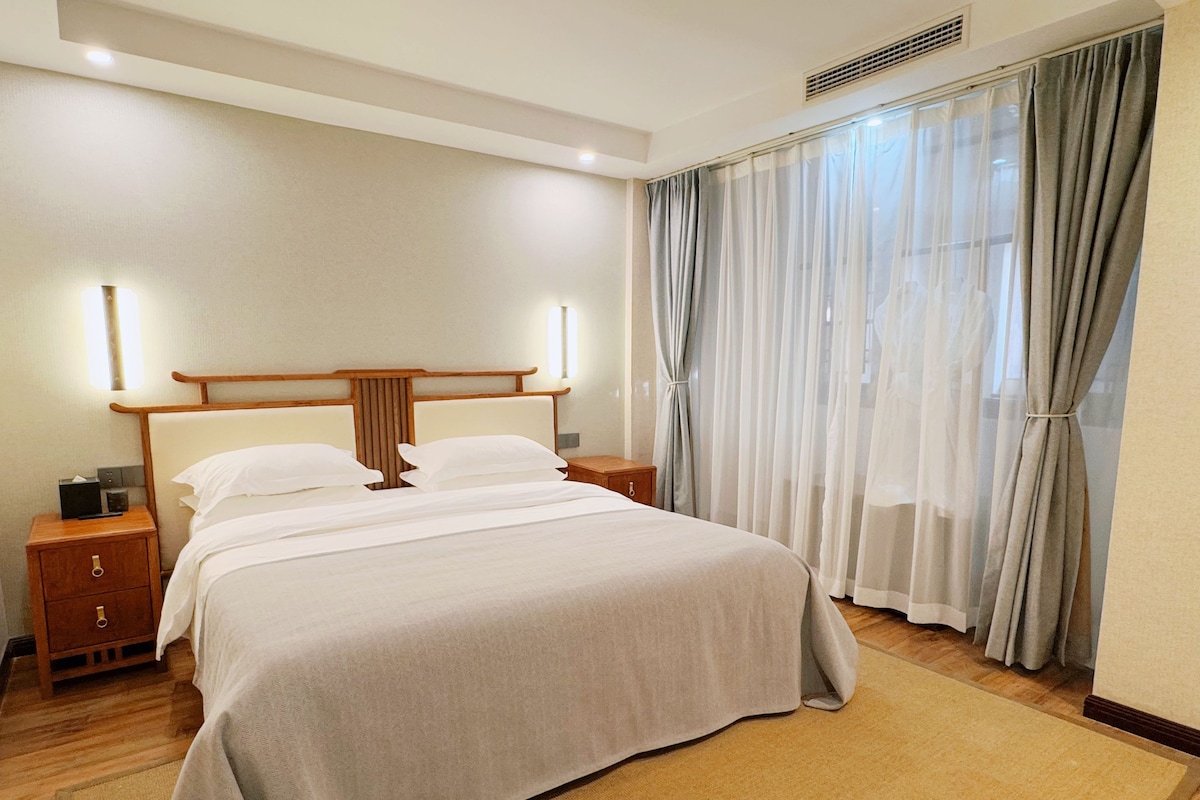 Une chambre d’hôtel avec un grand lit, une tête de lit en bois, des tables de chevet et une fenêtre avec des rideaux.