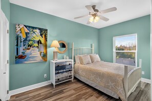 A bedroom with a bed, a nightstand, a ceiling fan, a window with blinds, and a wall painting. - Few Blocks to Beach • Pool Access + Golf Cart Zone | Beach Therapy (Port Aransas)
