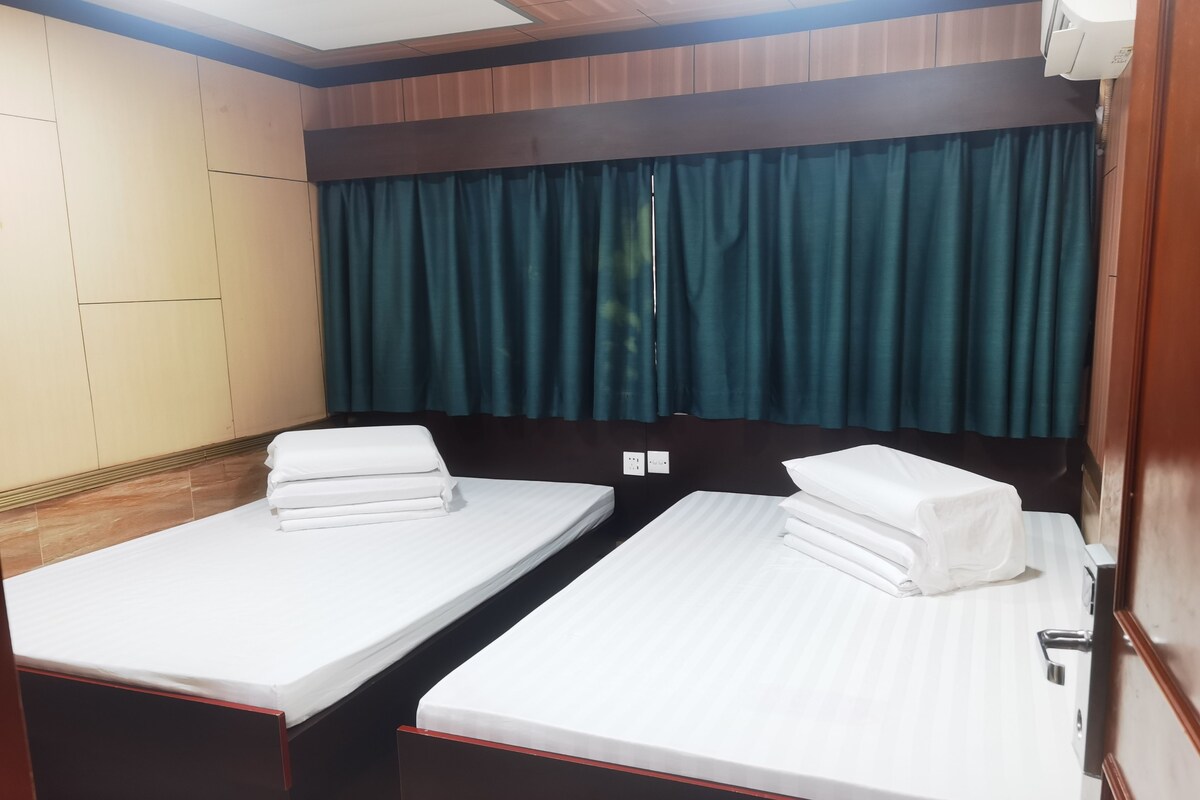 A hotel room with two single beds, a wooden headboard, a window with curtains, and a ceiling light.