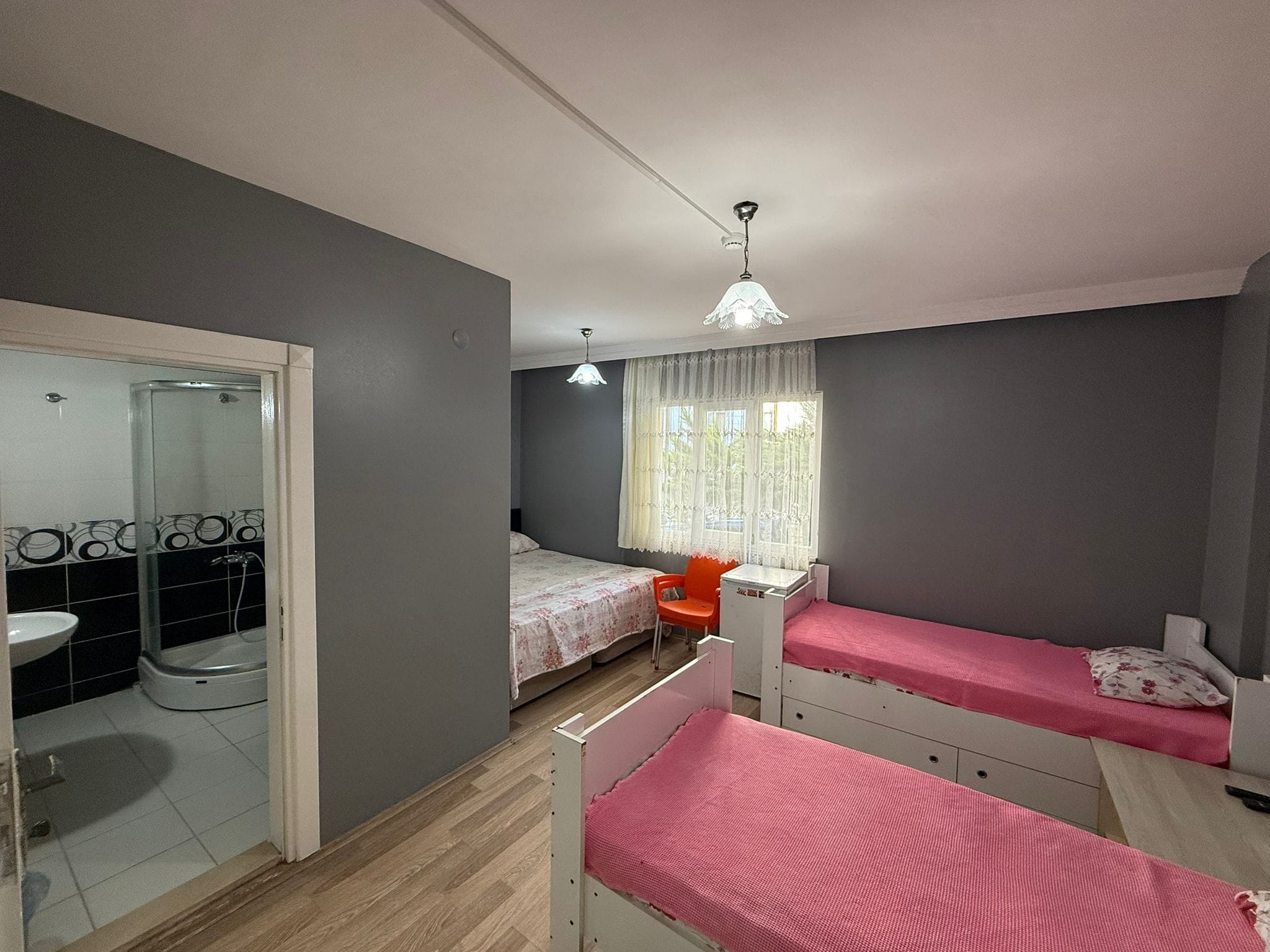 Habitación compacta con cama, escritorio, silla, ventana con cortinas y zona de ducha.