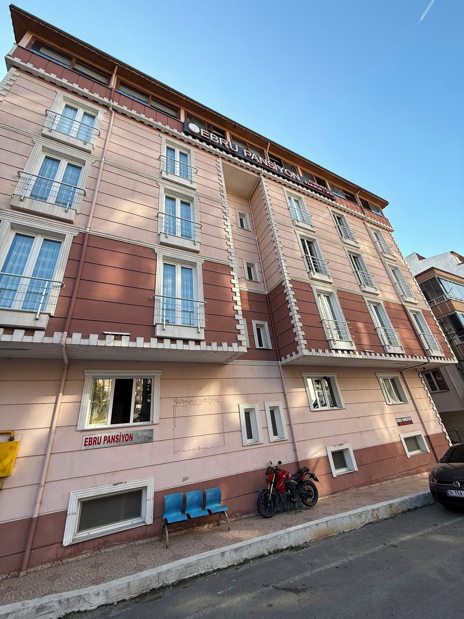 Un edificio de varios pisos con balcones, un letrero que dice 'EBRU PANSIYON' y una motocicleta estacionada.