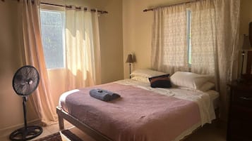 A bedroom with a bed, a standing fan, and a window with curtains.
