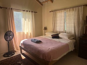 A bedroom with a bed, a standing fan, and a window with curtains. - Casa Baldoria Tropical Escape (Cabrera)