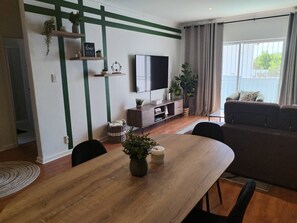 Een woonkamer met een houten eettafel, een bank, een televisie en een groot raam met gordijnen.