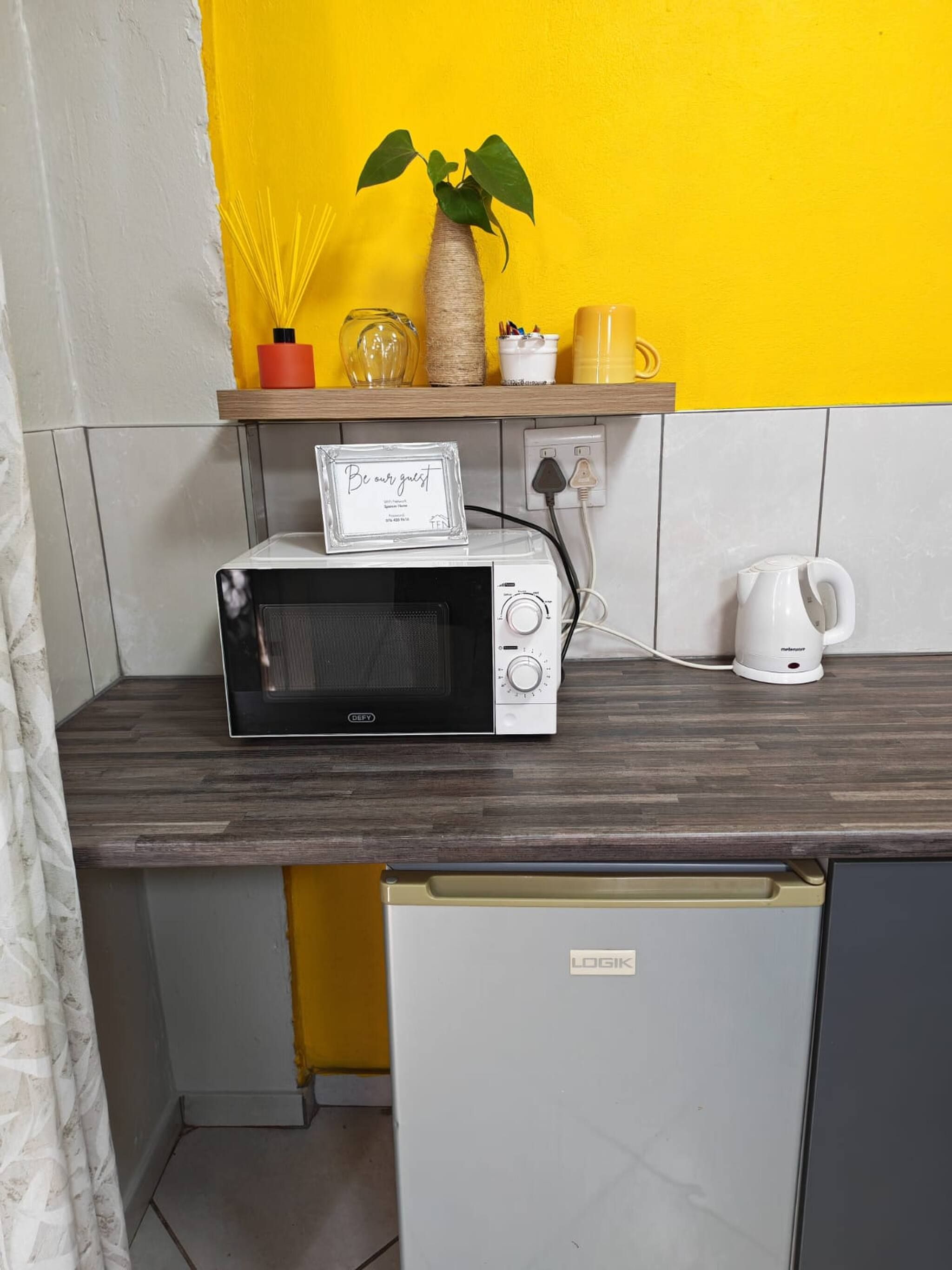 A kitchen with a microwave, a kettle, and a sign that says 'Be our guest.'