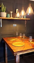A wooden dining table with four wine glasses and placemats, a potted plant, and pendant lighting.