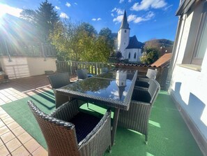 Une terrasse extérieure avec des meubles en osier et une table en verre, une vue sur une église et un espace recouvert d’un gazon synthétique vert.
