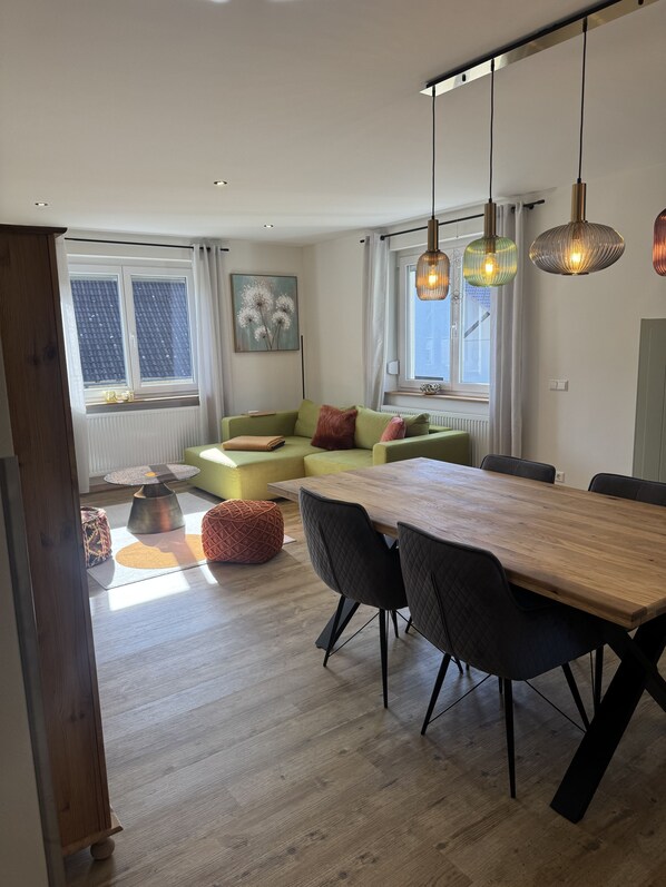 A modern living room with a wooden dining table, black chairs, a green sofa, and pendant lights. - Holiday Apartment 'Burghof' with Shared Garden and Wi-Fi (Leibertingen)