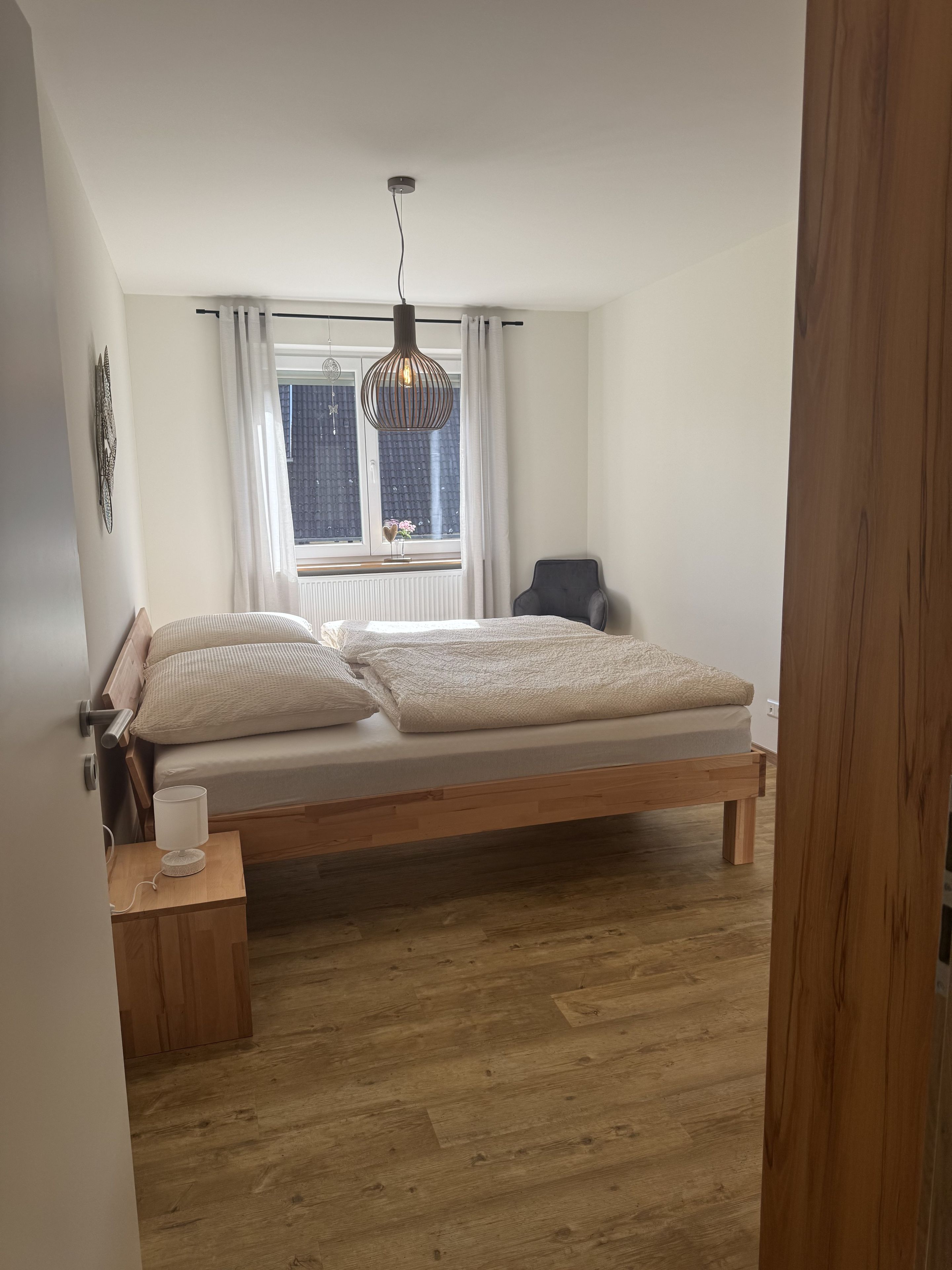 Et soveværelse med en træseng, et natbord med lampe, en stol, et vindue med gardiner og en hængende lysarmatur.
