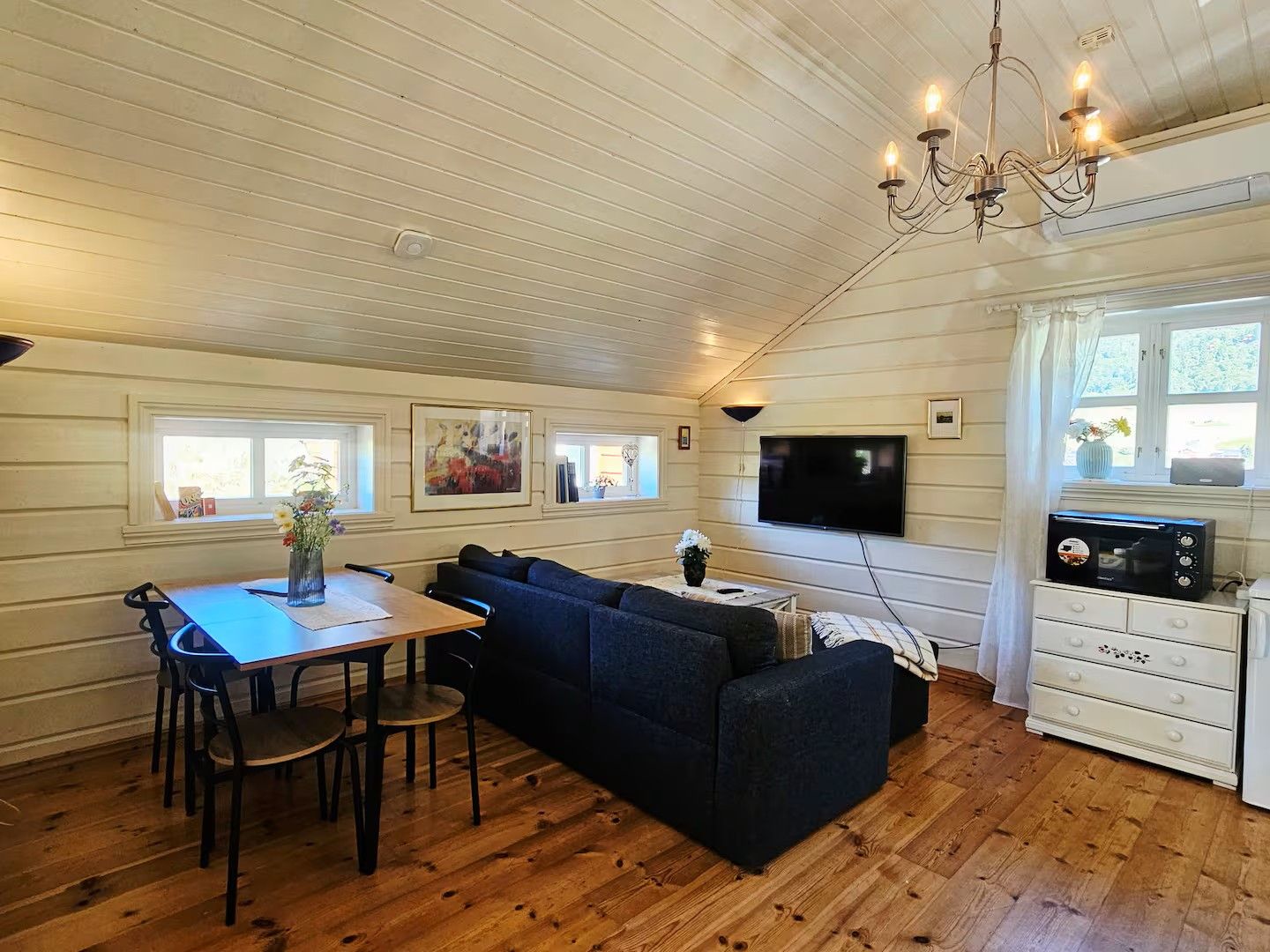Een gezellige woonkamer met een houten plafond, een televisie, een bank, een eettafel met stoelen, en een keuken met een magnetron.