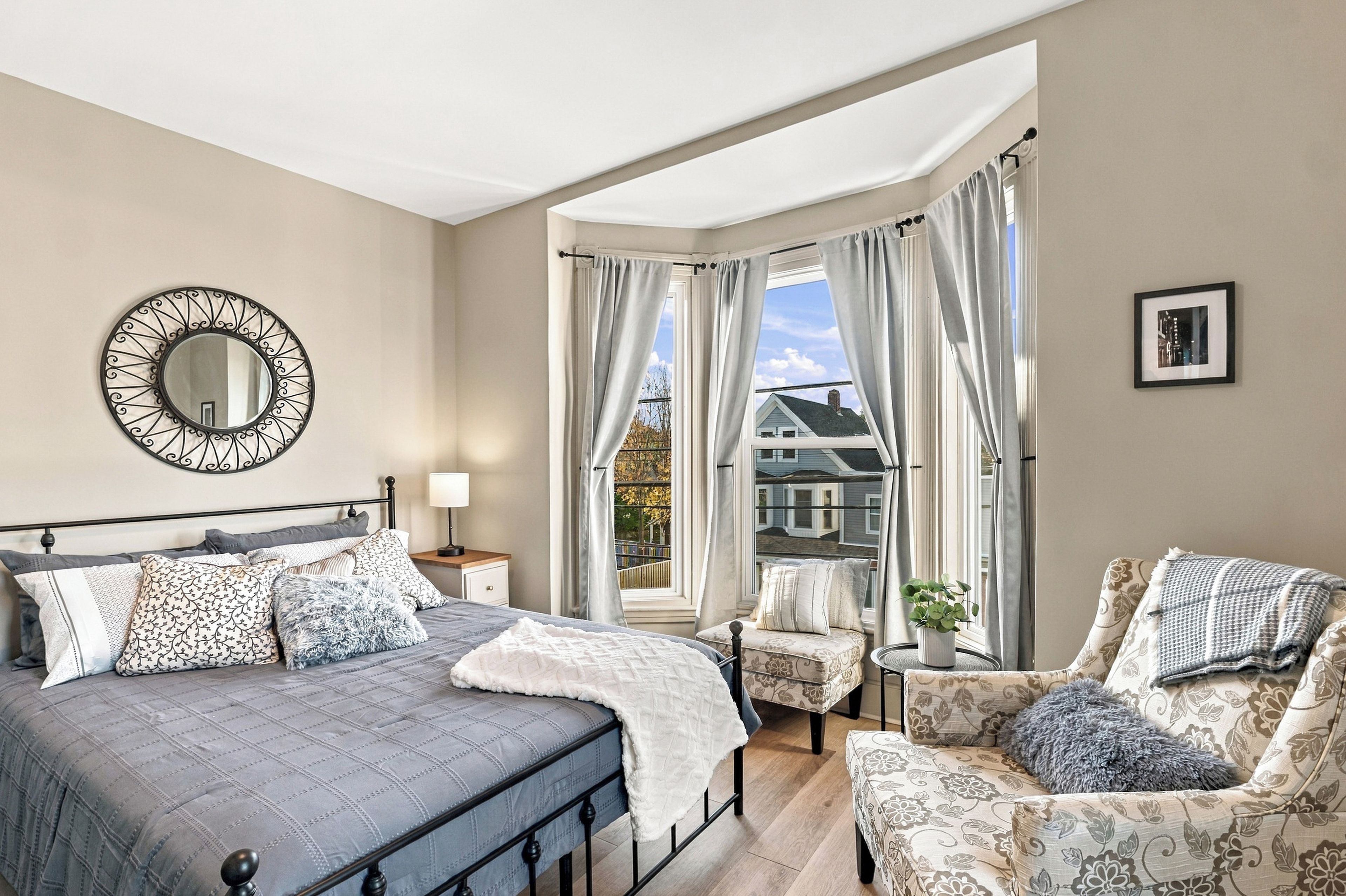 A bedroom with a bed, a chair, a small table, a mirror, and a window with curtains.
