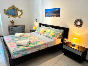 Una cama bien hecha con ropa de cama floral, una mesita de noche con una lámpara, un aparador, un espejo y un cuadro en la pared.