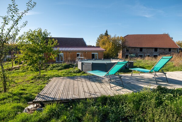 A wooden deck with two lounge chairs and a hot tub, surrounded by greenery and two buildings with distinct roofs.