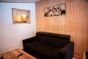 Un salon avec un canapé marron foncé, une table basse en bois et un téléviseur fixé au mur. Des tableaux encadrés ornent le mur.