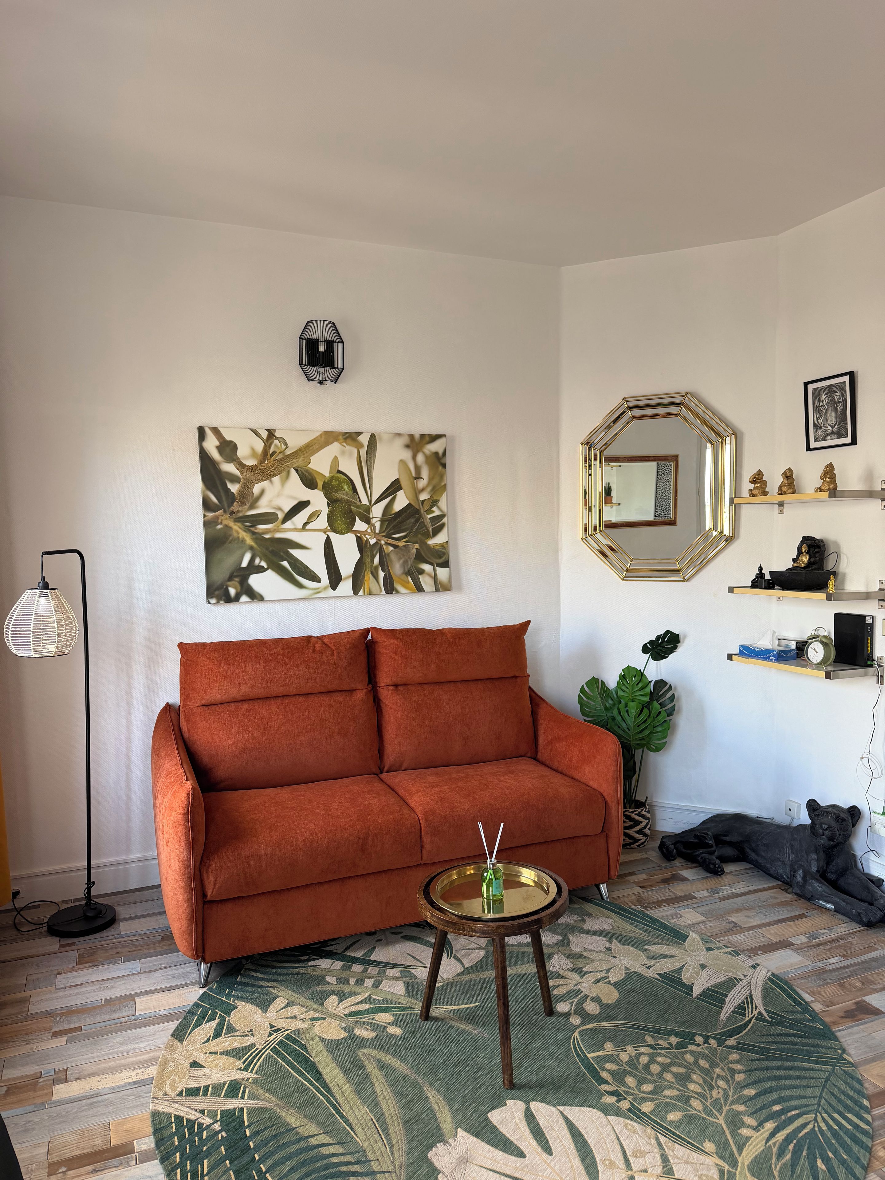 Uma sala de estar com sofá, mesa de centro redonda, uma grande obra de arte na parede, espelho e uma planta.