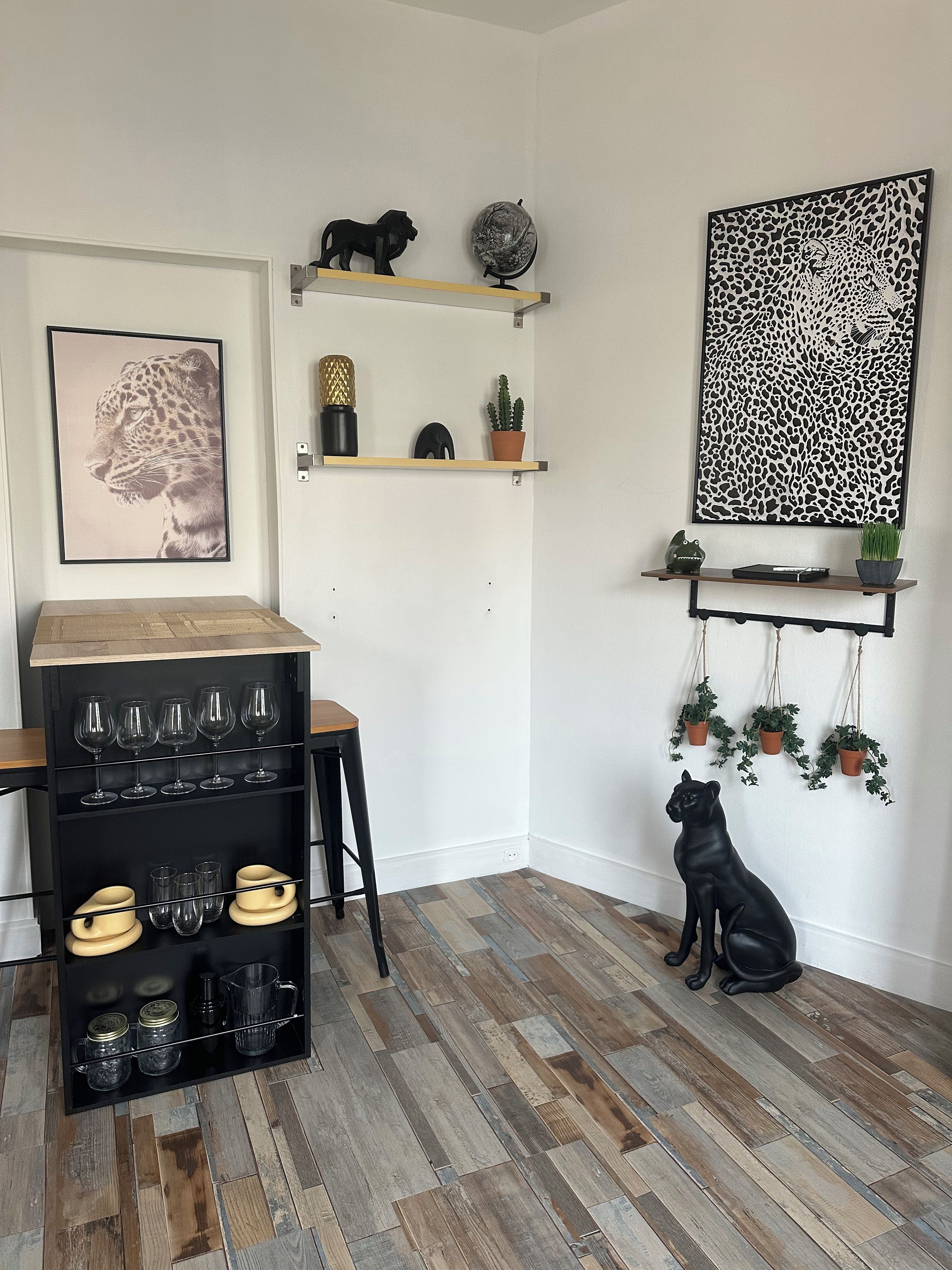 Quarto de canto com piso de madeira, um cão preto, comedouro preto para cão, prateleira preta com taças de vinho, obra de arte com estampa de leopardo emoldurada e um globo.