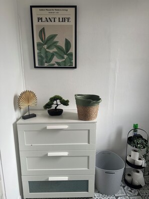 A white dresser with drawers, a framed plant art piece above it, a small bonsai tree, a woven basket, and a metal plant stand with rolled toilet paper. - New Listing (Vanves)