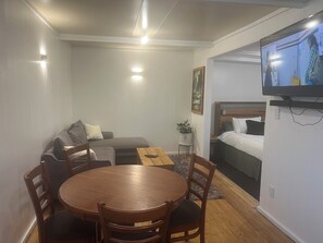 A compact living space with a sofa, a small dining table with chairs, a bed with a headboard, a television mounted on the wall, and a potted plant. - Mistypeaks Cottage (Okoki)